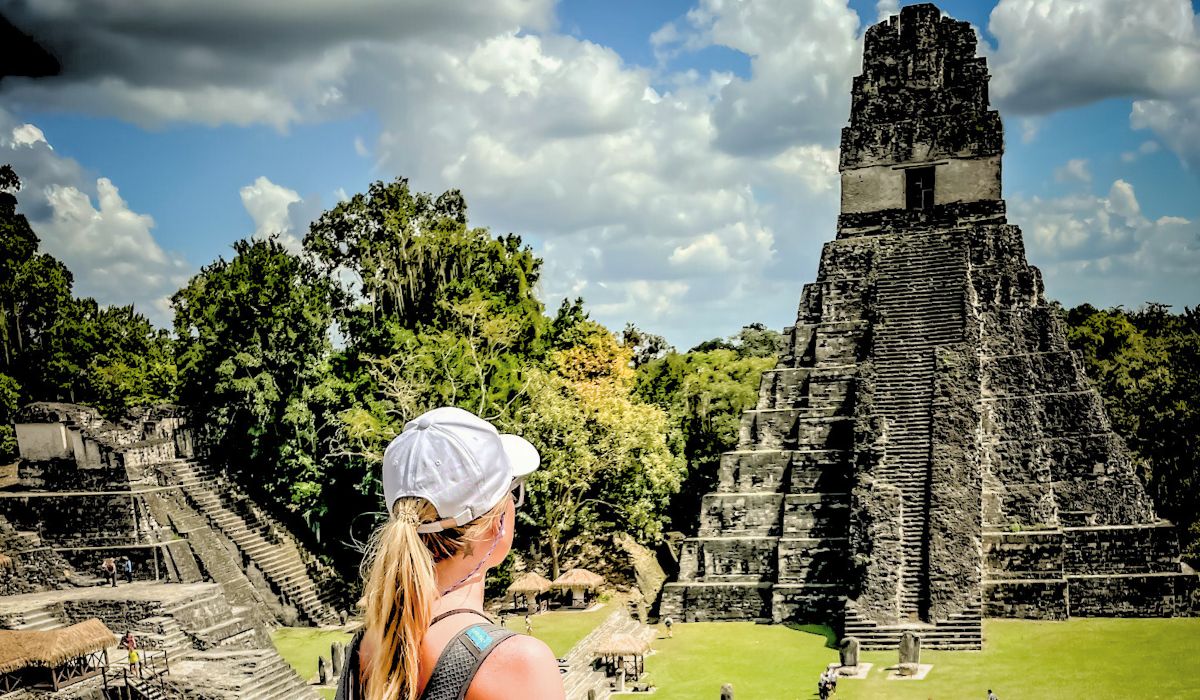 The image shows Kate, wearing a white cap and a backpack, standing and gazing at the majestic Temple I in Tikal, also known as the Temple of the Great Jaguar. The pyramid rises steeply in front of her, its steps leading up to the temple at the top. The surrounding area is filled with lush greenery and additional ruins, all under a partly cloudy sky.