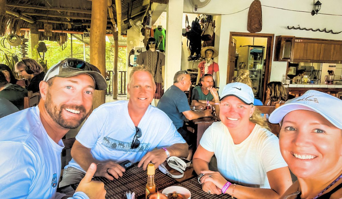 The image shows a group of four friends, including Kate and her husband, seated at a wooden table in a rustic, open-air restaurant. They are all smiling and appear to be enjoying their time together, with plates of food and bottles of hot sauce on the table. The background features other diners, tropical decor, and local crafts, creating a laid-back and authentic atmosphere that reflects the social and cultural experiences travelers might encounter on their journey from Belize to Tikal.