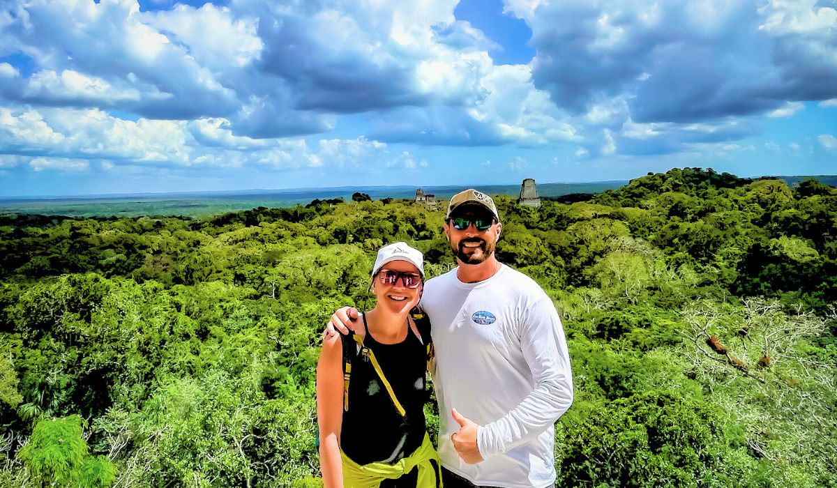 The image shows Kate and her husband standing together with a panoramic view of the Tikal ruins in the background. They are both smiling, with Kate wearing a white cap and sunglasses, and her husband giving a thumbs-up gesture. The vast jungle stretches out behind them, with ancient Mayan pyramids visible in the distance under a partly cloudy sky.