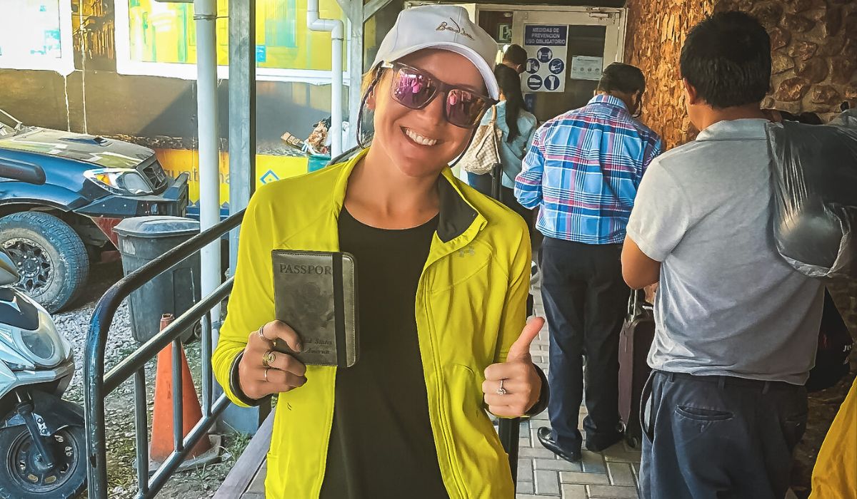 The image shows Kate standing in a line at what appears to be a border crossing or immigration checkpoint, holding her passport and giving a thumbs-up with a big smile. She is wearing a bright yellow jacket, a white cap, and sunglasses, ready for her journey. The background shows other travelers in line, with signs and vehicles visible, highlighting the travel process from Belize to Tikal