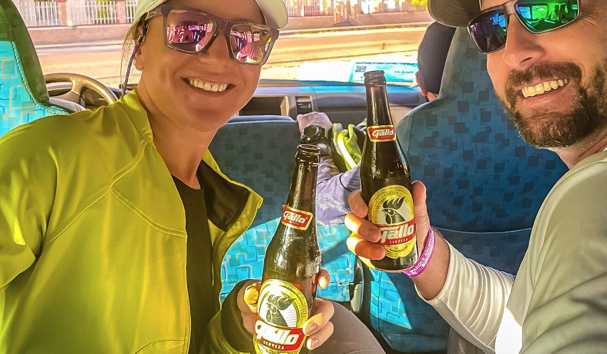 The image shows Kate and her husband sitting in the back of a tour bus, smiling and holding up bottles of Gallo beer as they enjoy a refreshing drink. They are both wearing sunglasses, with Kate in a bright yellow jacket, and their expressions suggest they're having a great time on their journey.