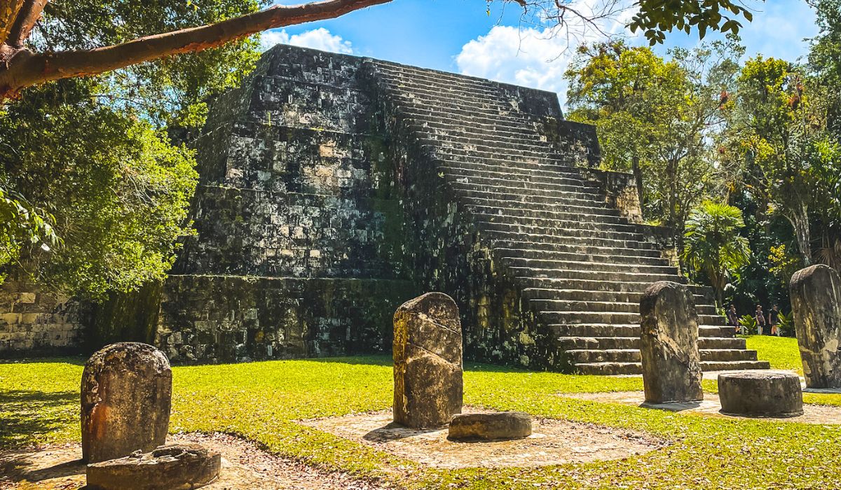 The image shows an ancient Mayan pyramid in Tikal, surrounded by stone stelae and altars, which are positioned in the foreground on a grassy area. The pyramid is steep, with weathered stone steps leading to the top, and is partially shaded by trees. The bright sky and sunlight filtering through the trees.