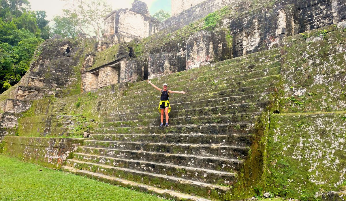 The image shows Kate standing on the moss-covered stone steps of an ancient Mayan structure in Tikal National Park. She is wearing a backpack and athletic attire, with her arms spread wide, looking joyful as she explores the ruins. The weathered stone of the pyramid-like structure, partially covered in greenery, highlights the historic and natural beauty of this popular destination.