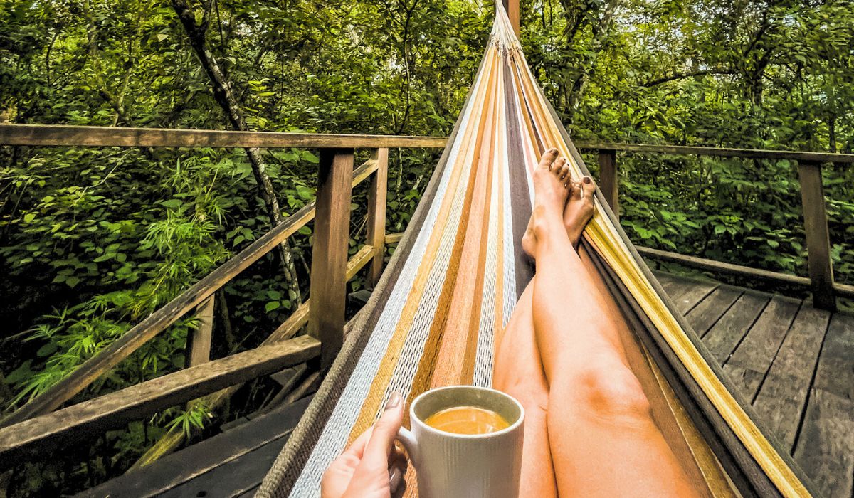 The image captures a serene moment of relaxation, with Kate lying in a striped hammock, holding a cup of coffee. The hammock is set on a wooden deck surrounded by dense, green foliage, creating a peaceful, nature-filled atmosphere.