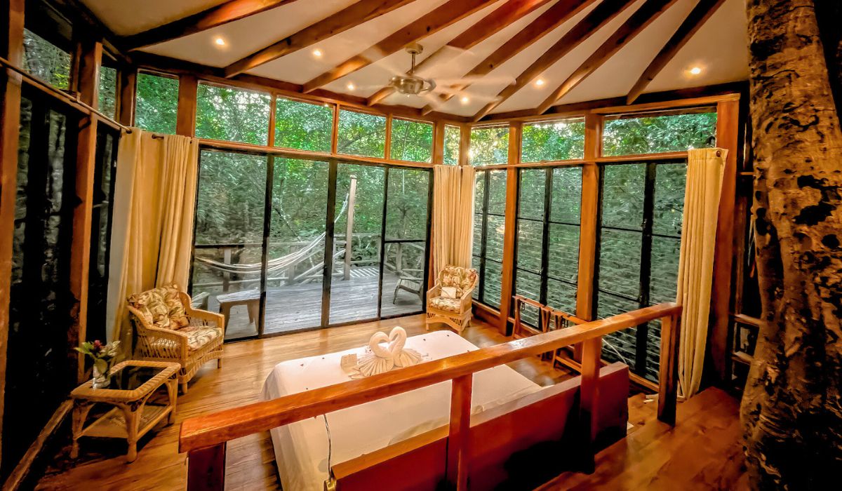 The image shows a cozy and inviting room with large windows that offer a panoramic view of the surrounding jungle. The room features a comfortable bed with towel art on it, wicker chairs, and a wooden deck outside, where a hammock is strung up for relaxation. The natural light flooding through the windows and the tree-trunk beam inside the room emphasize the close connection with nature, making it an ideal retreat for those traveling from Belize to Tikal.