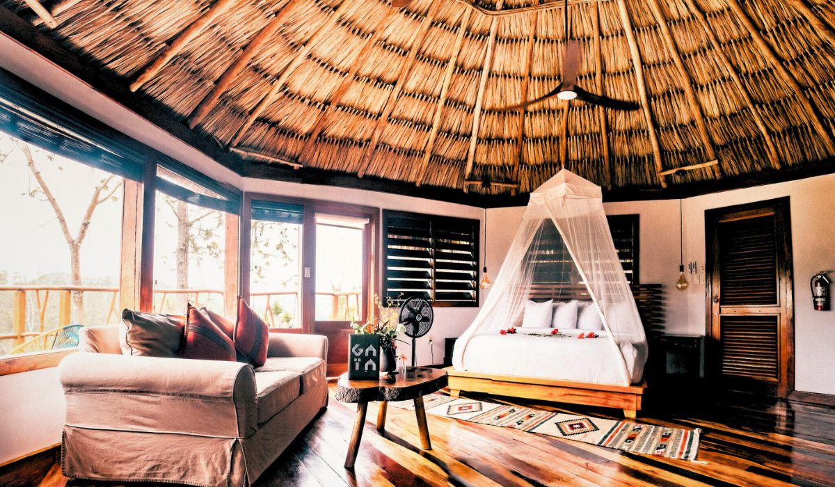 The image shows a beautifully designed interior of a thatched-roof bungalow, featuring a spacious bedroom with a large bed draped in a white mosquito net. The room is warmly lit and includes a comfortable sofa, wooden furniture, and decorative rugs, all of which create a cozy, inviting atmosphere. The large windows allow for plenty of natural light and offer views of the surrounding natural landscape.