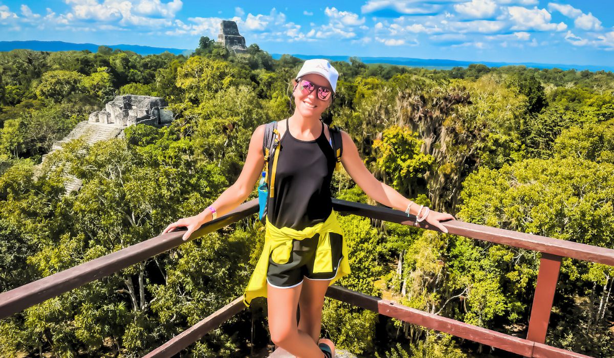 The image shows Kate standing on a wooden observation deck with a stunning panoramic view of the Tikal ruins in the background. She is smiling, dressed in athletic attire with a yellow jacket tied around her waist, and wearing a white cap and sunglasses. The lush green jungle spreads out behind her, with ancient Mayan pyramids peeking through the treetops, illustrating the grandeur of Tikal, a must-see destination.