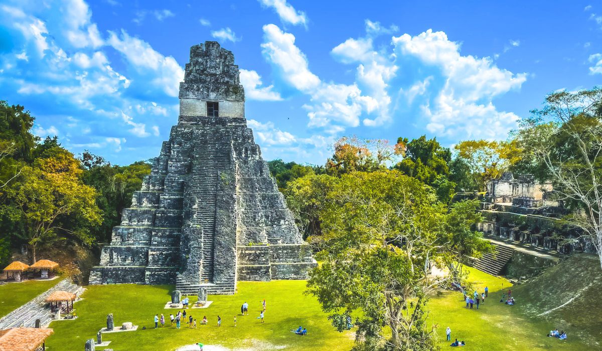 The image shows the iconic Temple I at Tikal, an ancient Mayan ruin located in a lush, green jungle setting. The stone pyramid rises steeply, surrounded by scattered tourists exploring the historical site. The bright blue sky with scattered clouds adds to the beauty of the scene. The image captures the majesty of this Guatemalan archaeological wonder, which is a popular day trip destination for visitors traveling from Belize to Tikal.