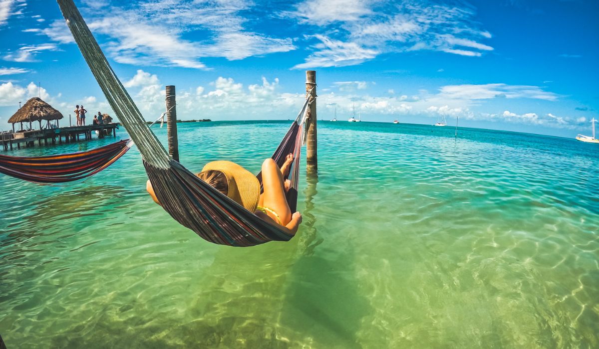 Kate relaxes in a hammock suspended over clear, shallow waters, with a wide-brimmed hat shading their face. The idyllic scene, set against a backdrop of turquoise seas and blue skies, epitomizes the tranquil, laid-back atmosphere of a Belizean beach getaway.