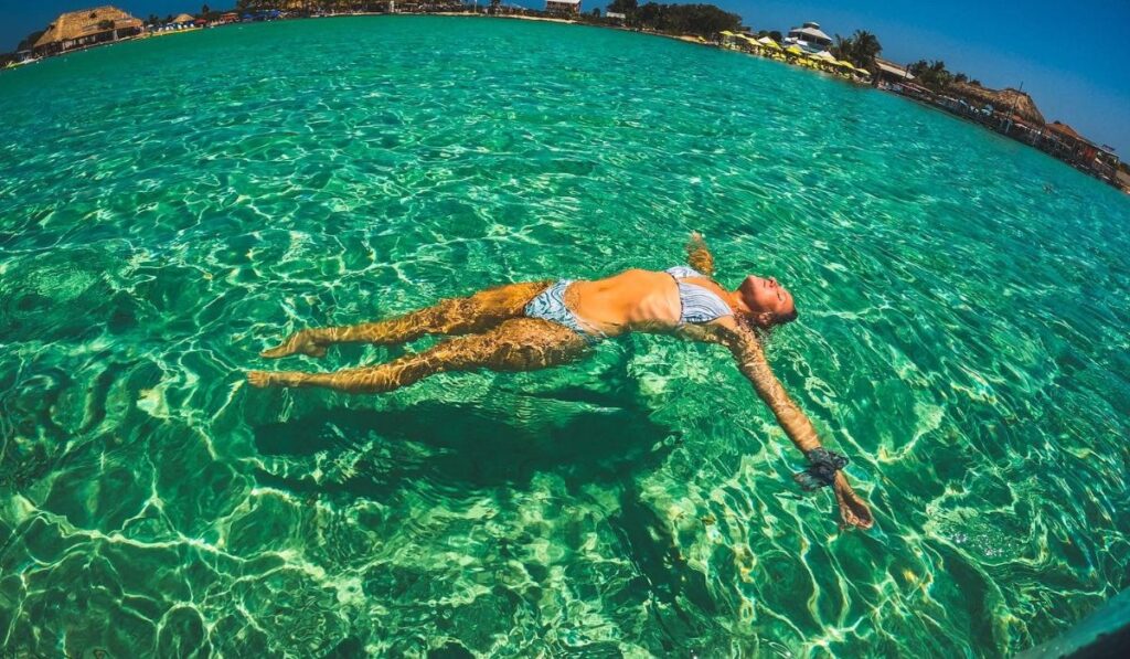 A woman floats effortlessly on her back in the crystal-clear turquoise waters of Belize, basking in the sun with her arms outstretched. The vibrant water and distant shoreline create a serene, relaxing atmosphere.