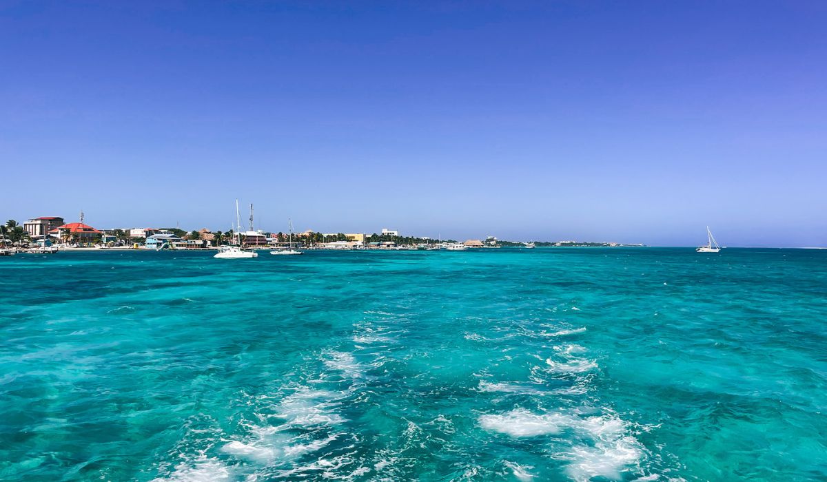 A view from the water of a coastal town in Belize, with turquoise waves trailing behind a boat. The shoreline is lined with buildings and boats anchored in the harbor, all under a clear, bright blue sky, highlighting the scenic and vibrant coastal life of Belize.