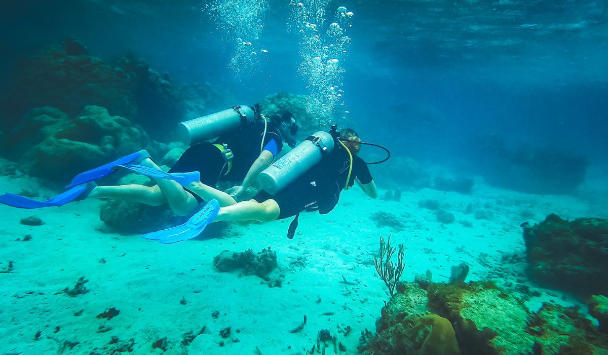 Two scuba divers explore the underwater world in Belize, swimming side by side above the sandy ocean floor, surrounded by coral formations. Bubbles rise as they move through the clear blue water.