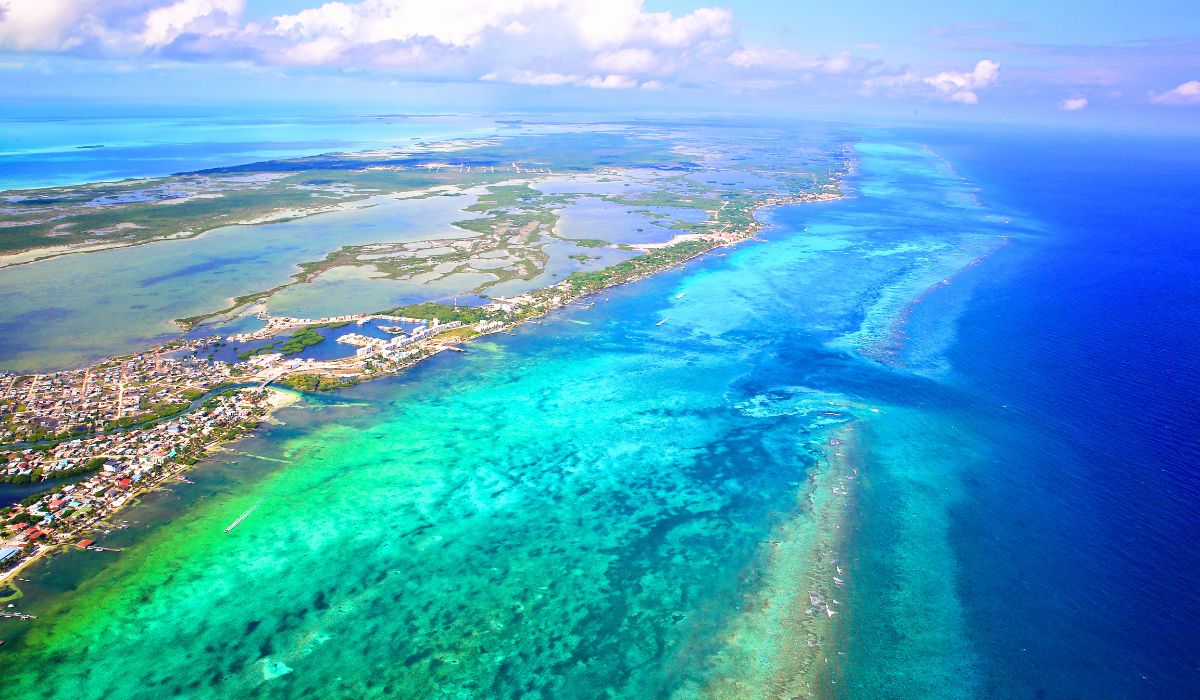 An aerial view of Ambergris Caye, Belize, showing the vibrant turquoise waters of the Caribbean Sea meeting the coastline. The island's mix of urban areas, green spaces, and expansive reef systems are visible.