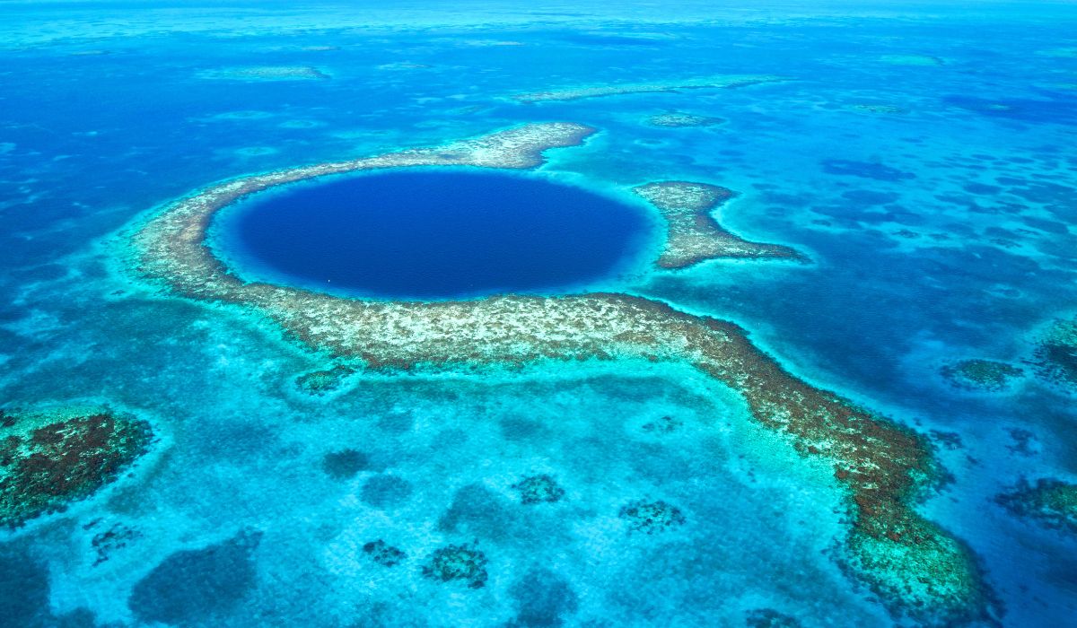 An aerial view of the Great Blue Hole, a massive marine sinkhole surrounded by turquoise waters and coral reefs off the coast of Belize. The deep blue circular formation stands out vividly against the lighter blue and green hues of the surrounding ocean, highlighting one of Belize's most famous natural wonders.