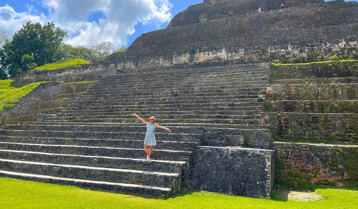 Kate stands at the base of an ancient Mayan pyramid at Xunantunich in Belize, arms outstretched, surrounded by the massive stone steps of the ruins. The bright blue sky and lush greenery frame the historical site.