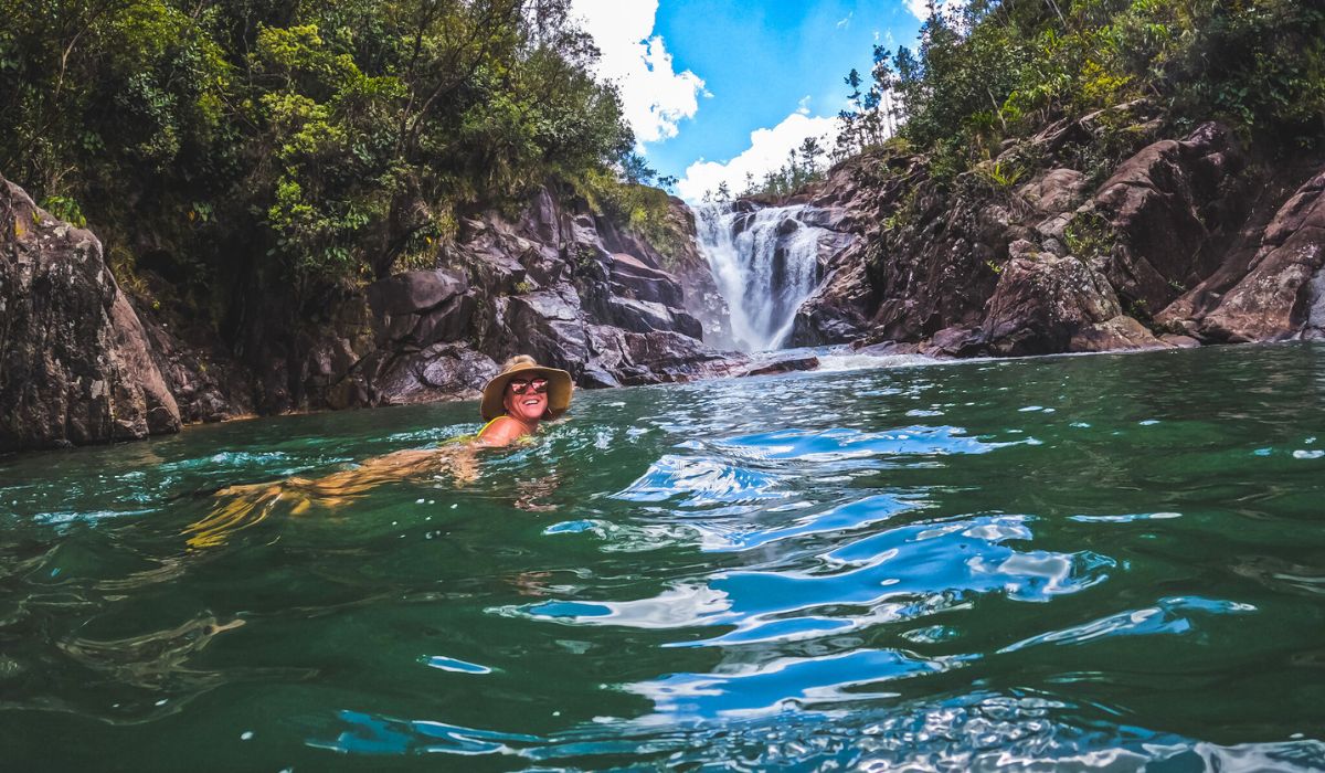 Kate, wearing a sun hat, enjoys a swim in a natural pool at the base of a waterfall in Belize, surrounded by lush green vegetation and rocky cliffs. The clear blue sky and cascading water create a serene and adventurous atmosphere, highlighting the beauty of Belize's natural landscapes.