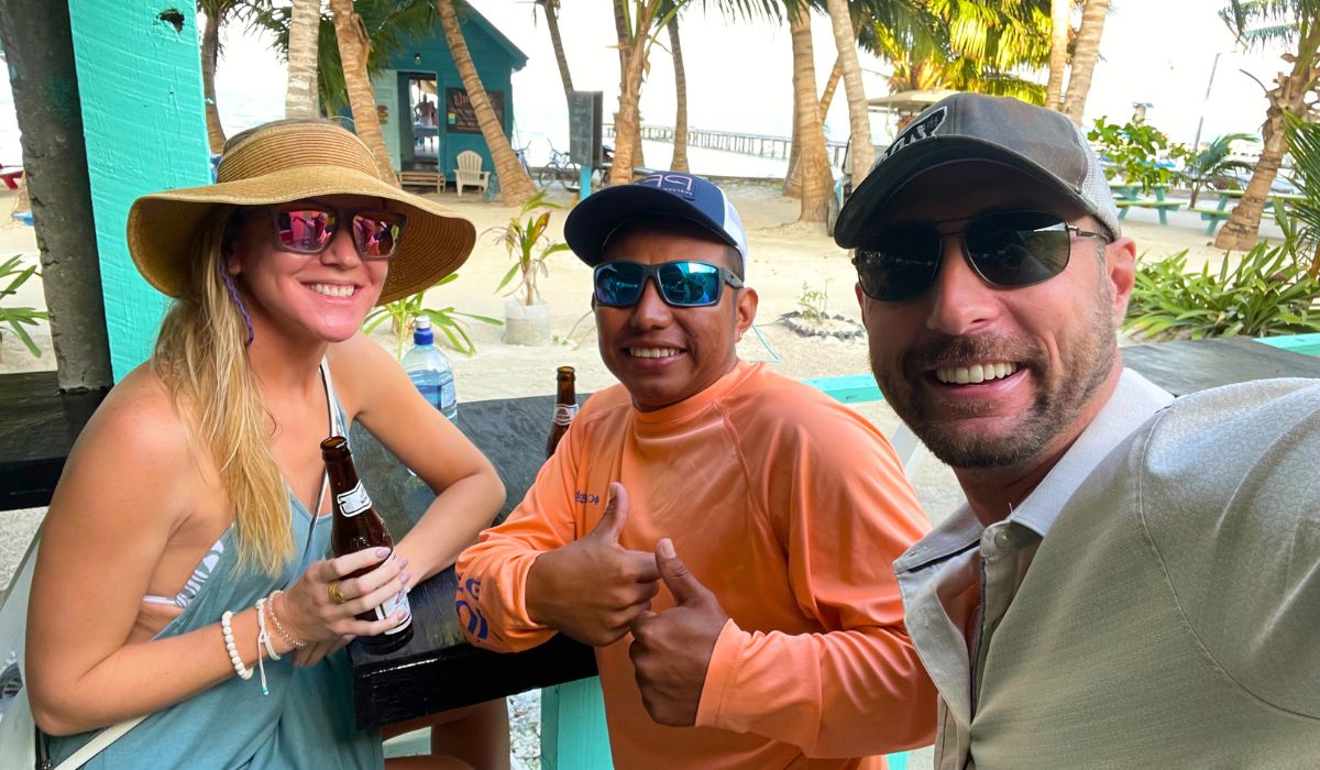 Kate and her husband, and a local, enjoy drinks together at a beachfront bar in Belize. The group is all smiles under the shade of palm trees, capturing the friendly and relaxed atmosphere among Belize locals.