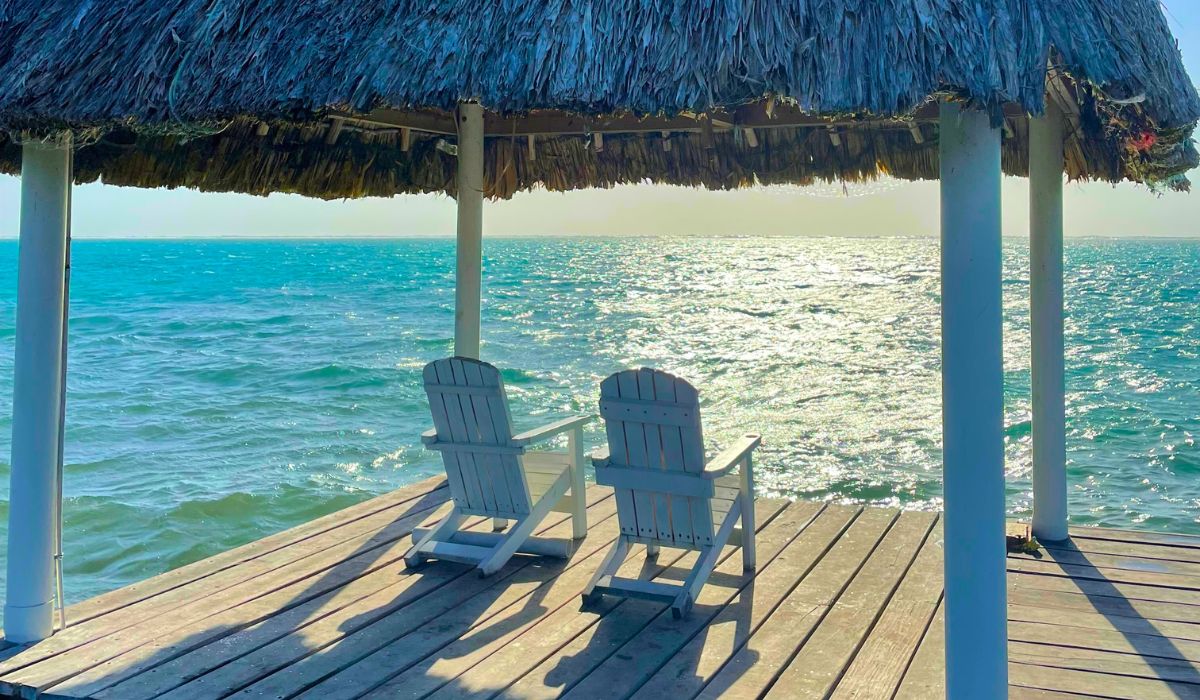 Two white wooden chairs sit under a thatched-roof palapa on a wooden deck overlooking the calm, turquoise waters of the Caribbean Sea in Belize. This serene and picturesque setting underscores the question, "Is Belize worth visiting?" by showcasing the tranquil beauty and inviting atmosphere of this tropical paradise.