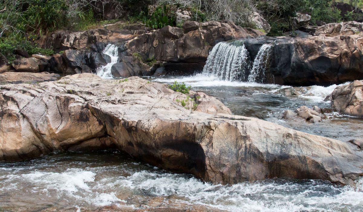 The image depicts a serene scene of a small waterfall cascading over rocky terrain into a clear, flowing river. Surrounded by lush greenery and rugged rocks, this natural landscape captures the tranquil beauty of a hidden spot in San Ignacio, Belize.