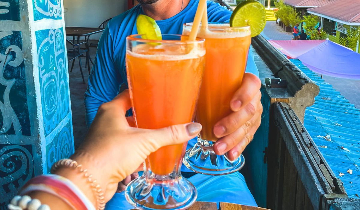 Two people toasting with tall glasses of bright orange drinks garnished with lime slices at a café overlooking the San Ignacio town square. The background shows colorful rooftops and outdoor seating areas, capturing a lively and cheerful atmosphere.