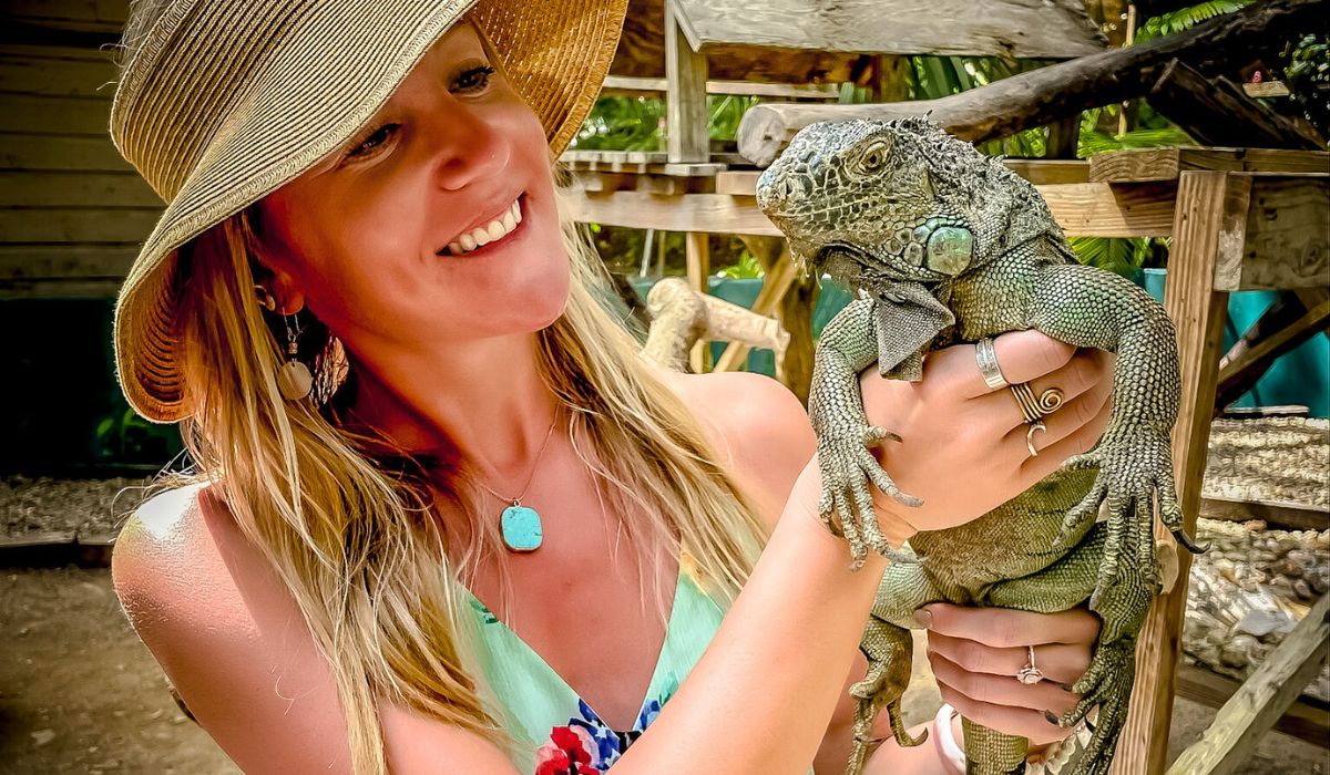 Kate, smiling, wearing a straw hat, holds a large iguana at the Green Iguana Conservation Project in San Ignacio, Belize. The background shows a wooden enclosure and greenery, highlighting the natural habitat of the iguanas.