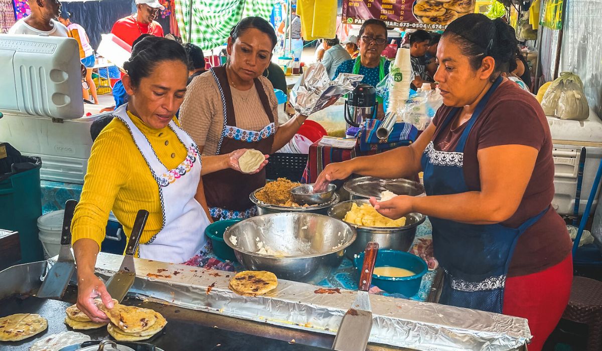 Women preparing and cooking traditional food at a bustling market stall in San Ignacio, Belize. They are working with dough and various fillings, creating handmade tortillas or pupusas on a large griddle. The vibrant market atmosphere is filled with colorful fabrics and busy vendors