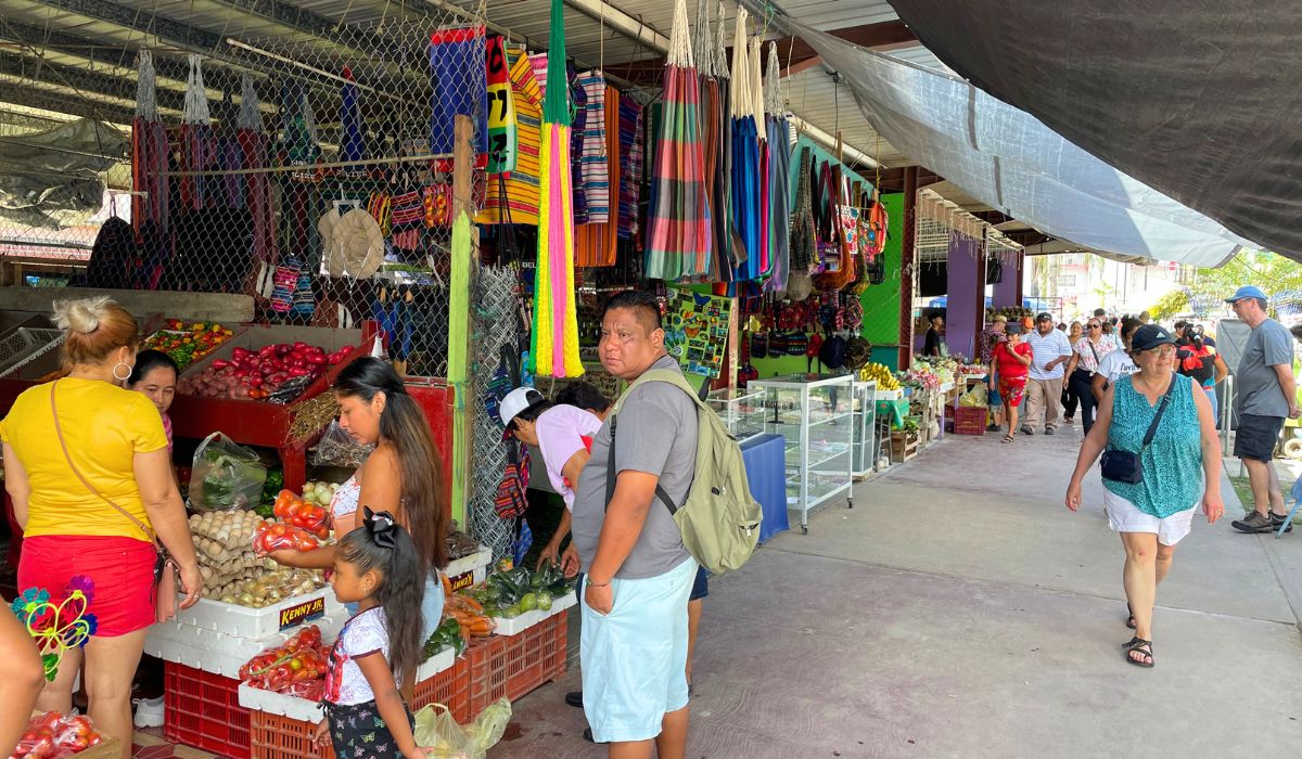 Shoppers browse through a vibrant local market in San Ignacio, Belize, featuring fresh produce and colorful textiles. The stalls display a variety of fruits, vegetables, and handcrafted goods, with people engaging in lively conversations and transactions.