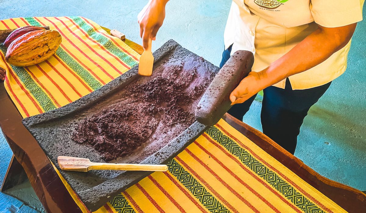 A person demonstrating traditional chocolate making by grinding cacao beans on a stone metate at a workshop in San Ignacio, Belize. The table is adorned with a colorful striped cloth and a yellow cacao pod.