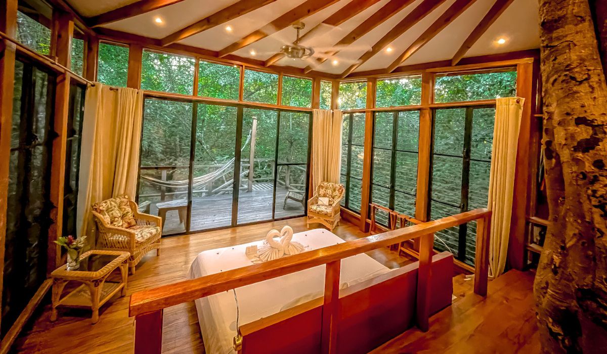 A cozy and elegant room at Vanilla Hills Lodge, featuring large windows that offer a panoramic view of the lush jungle outside. The room includes a comfortable bed with swan towel decorations, wicker furniture, and a private deck with a hammock.