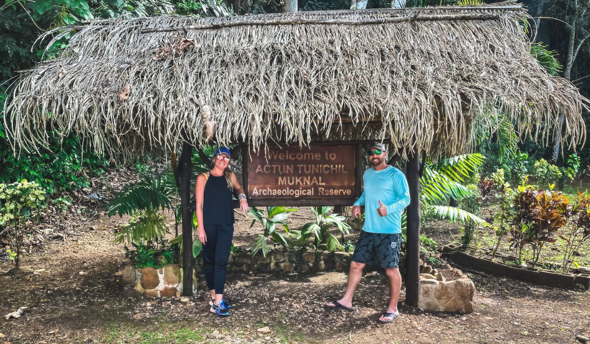 The image shows Kate and her husband standing under a thatched-roof sign that reads "Welcome to Actun Tunichil Muknal Archaeological Reserve." They are smiling and posing for the photo, surrounded by lush greenery, indicating the entrance to a significant archaeological site in San Ignacio, Belize.