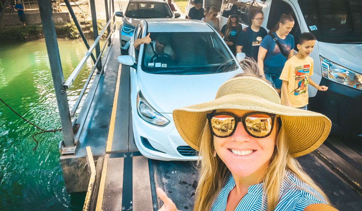 Kate in a straw hat and sunglasses takes a selfie while on a small car ferry in San Ignacio, Belize. Behind her, cars and people are visible on the ferry, with the green water of a river surrounding them.