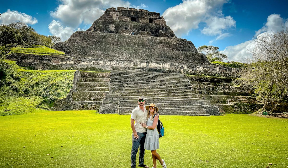 Kate and her husband pose in front of the majestic El Castillo pyramid at the Xunantunich Mayan ruins near San Ignacio, Belize. The massive stone structure rises impressively against a backdrop of blue sky and scattered clouds, with the couple standing on the bright green grass at its base.