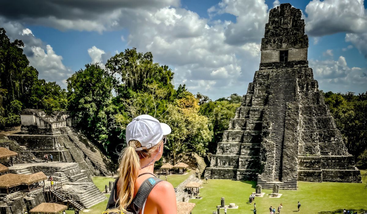 The image shows Kate with a backpack and a white cap looking at the iconic Temple I, also known as the Temple of the Great Jaguar, in the ancient Mayan city of Tikal, Guatemala. The pyramid stands tall amidst lush greenery and other ruins, illustrating the grandeur and historical significance of the site.