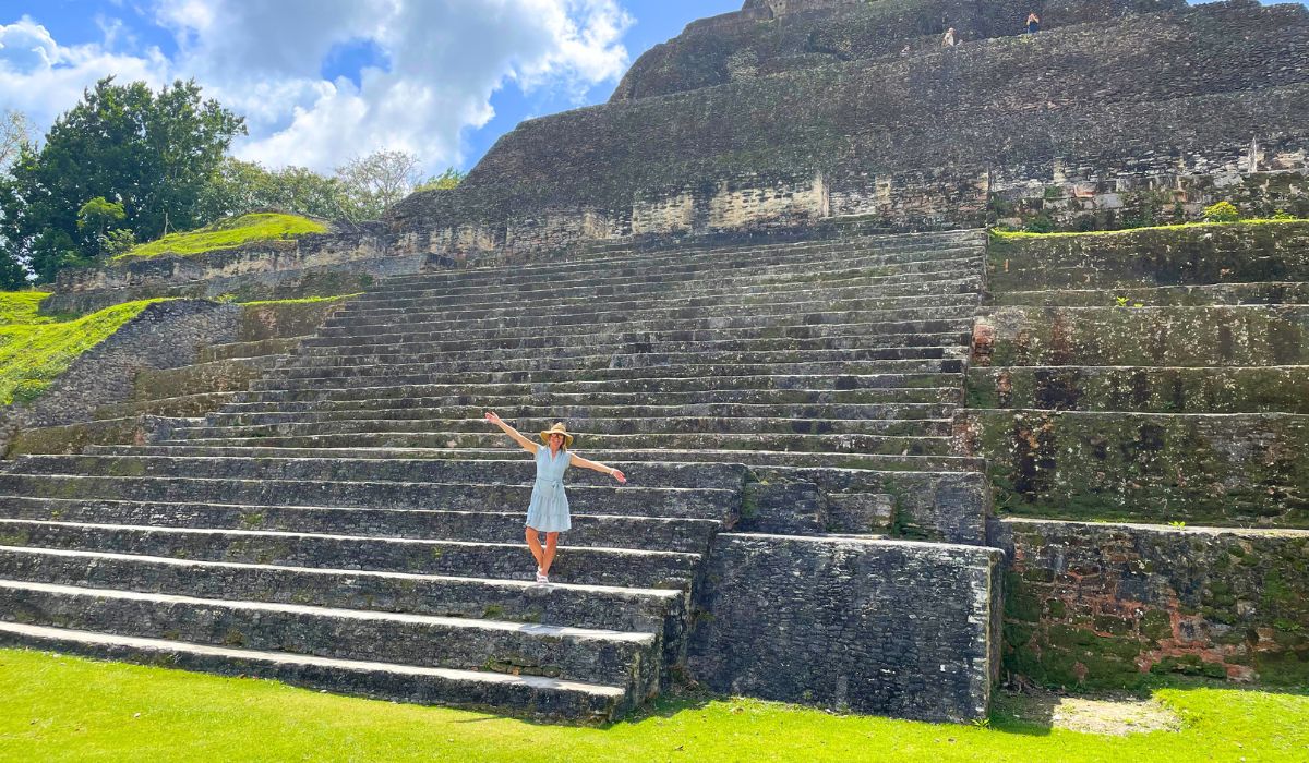 The image shows Kate standing with her arms outstretched on the steps of an ancient Mayan pyramid in Xunantunich. She is wearing a light blue dress and a hat, enjoying the sunny day at the impressive archaeological site, which is surrounded by lush greenery and clear skies in San Ignacio, Belize.