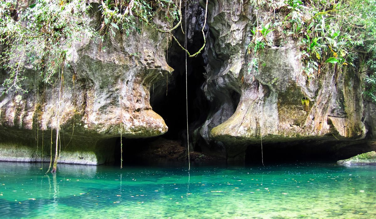 The image shows the entrance to a cave with lush greenery hanging over its rocky opening and clear turquoise water at its base. This scenic spot is ideal for cave tubing, a popular adventure activity in San Ignacio, Belize.