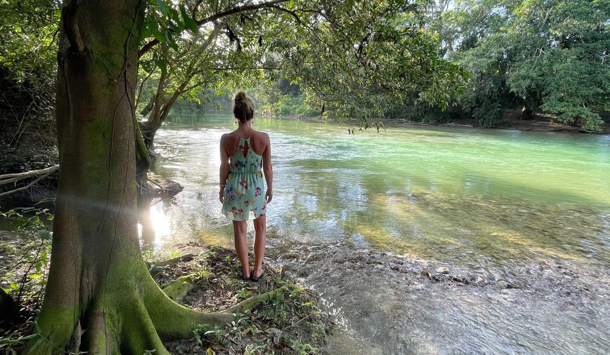 The image shows Kate in a floral dress standing on the bank of the Macal River, surrounded by lush greenery and illuminated by soft sunlight filtering through the trees. The clear, tranquil waters of the river create a serene and picturesque setting in San Ignacio, Belize.