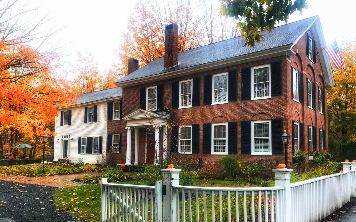 Charming colonial-style brick house in Woodstock, Vermont, New England, surrounded by vibrant fall foliage, with small pumpkins adorning the front yard.