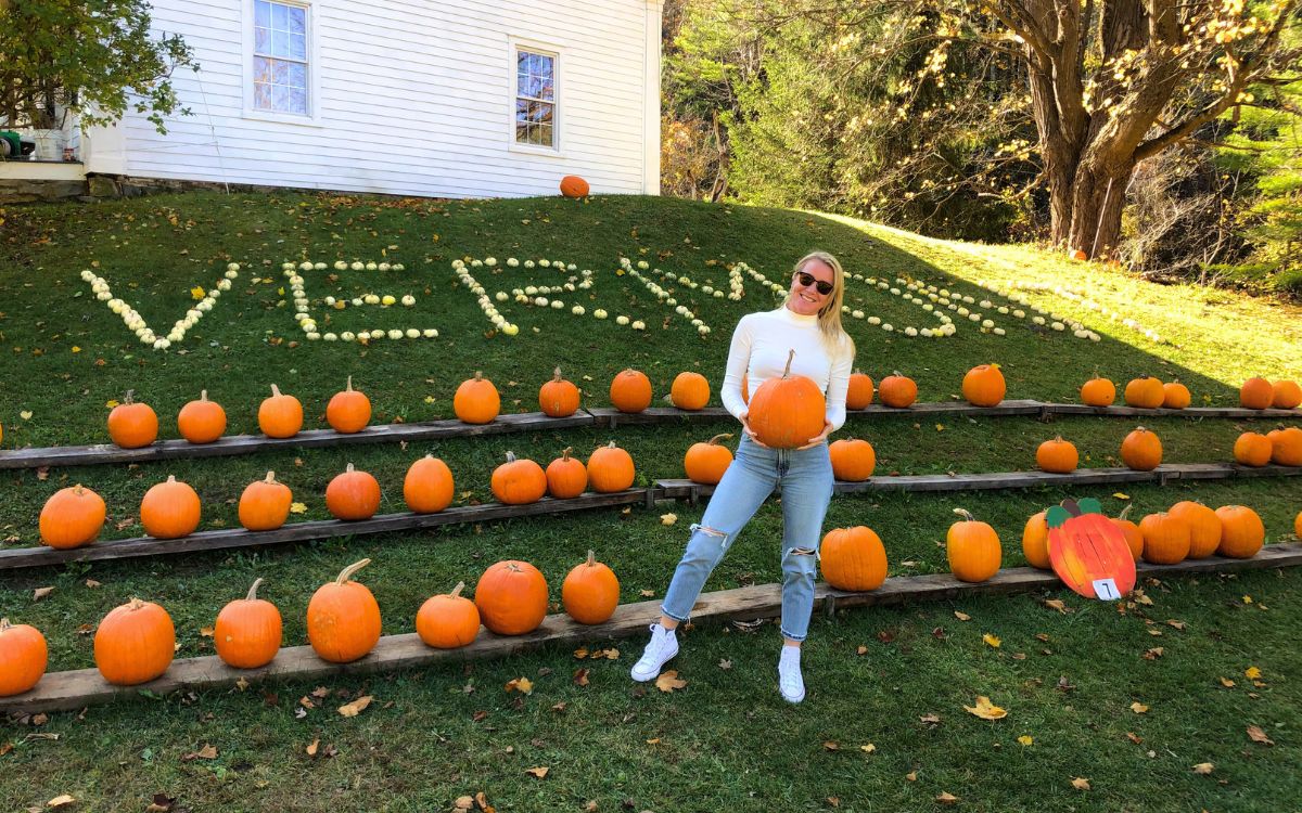 Kate smiling while holding a large pumpkin, standing in front of a display of pumpkins with the word 'Vermont' spelled out on the hillside behind her.