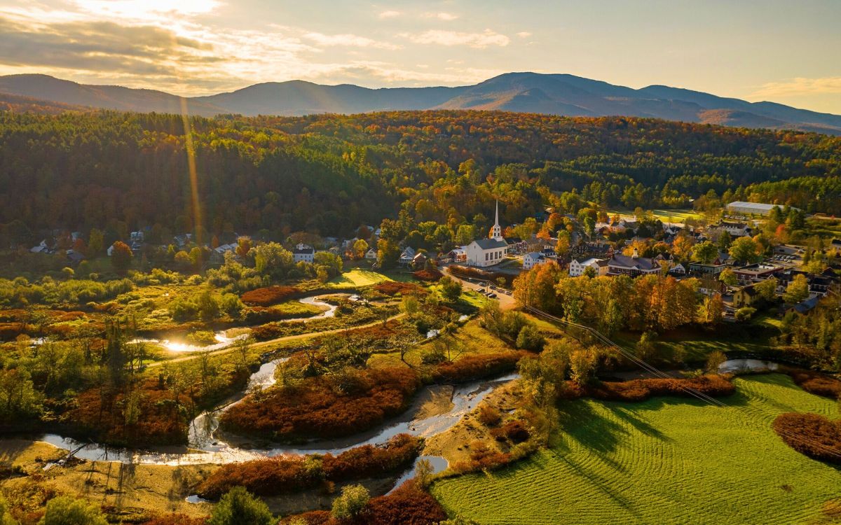 Aerial view of Stowe, Vermont, a picturesque New England village nestled among vibrant autumn foliage, with a winding river, green fields, and mountains in the background during sunset.