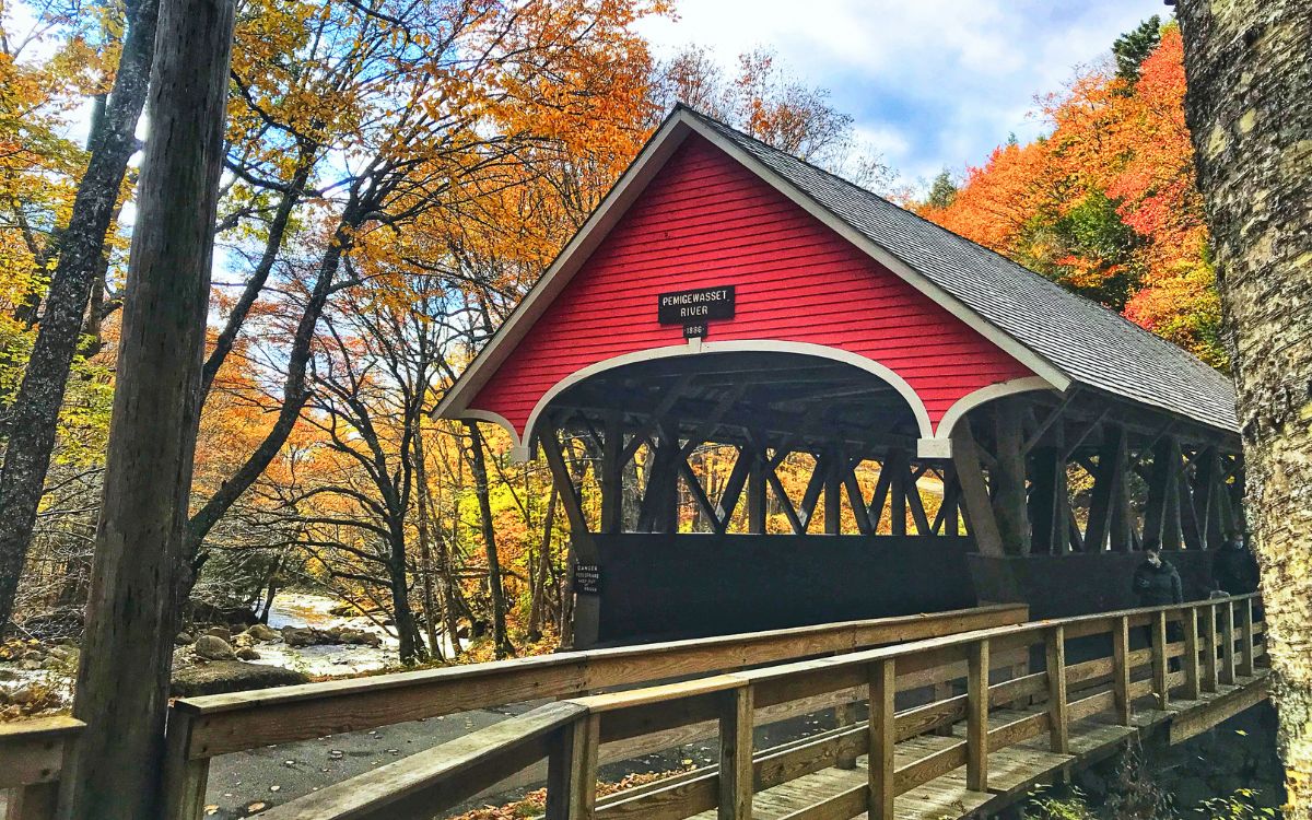 Red covered bridge over the Pemigewasset River in New England, surrounded by colorful autumn trees, with a wooden walkway leading to the bridge.