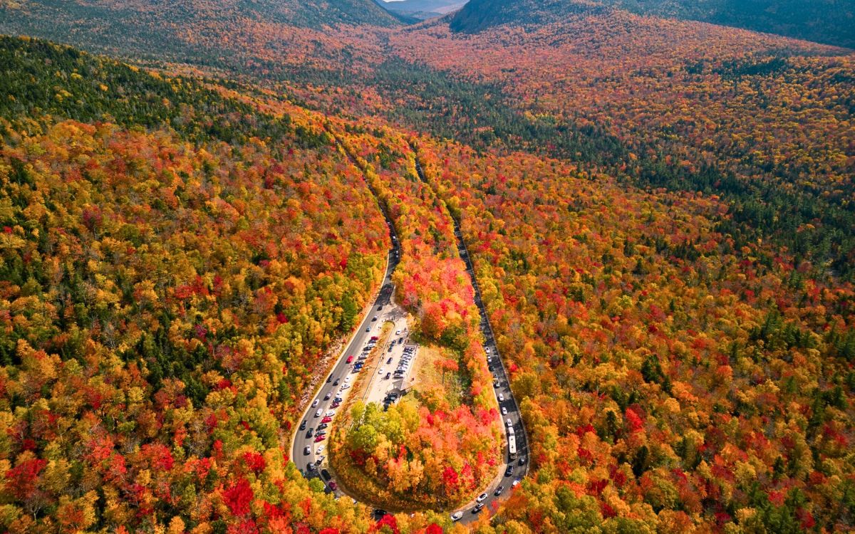 Aerial view of the winding Kancamagus highway surrounded by dense forest covered in vibrant fall colors, with cars parked along the road, capturing the autumn beauty in New England.