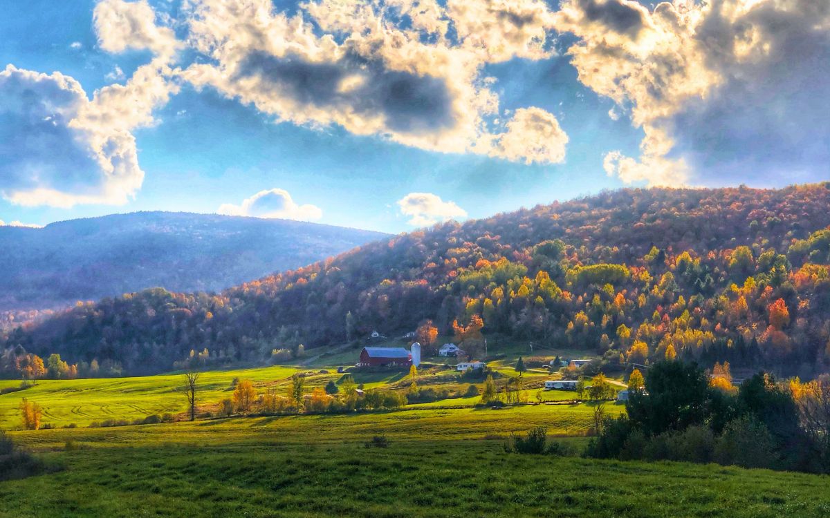 Scenic view of a rural farm nestled in a valley in Vermont, surrounded by colorful autumn trees on rolling hills under a dramatic sky in New England.