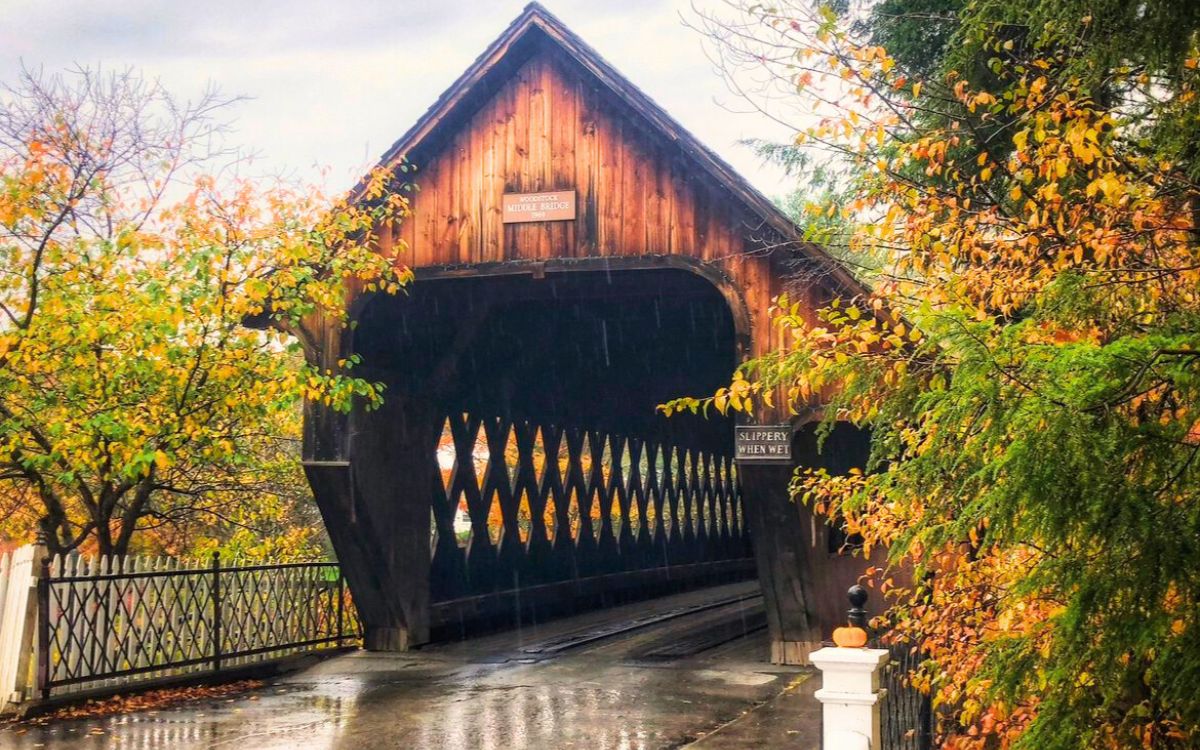 Rain-soaked covered bridge in New England, surrounded by autumn trees with colorful leaves, and a sign reading 'Slippery When Wet' at the entrance.