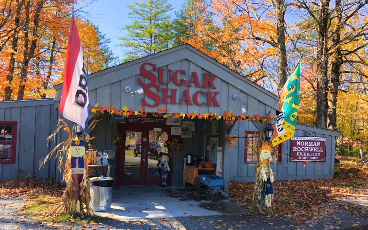 Entrance of the Sugar Shack gift shop decorated for fall, with colorful foliage, scarecrows, and signs promoting a Norman Rockwell exhibition in New England.