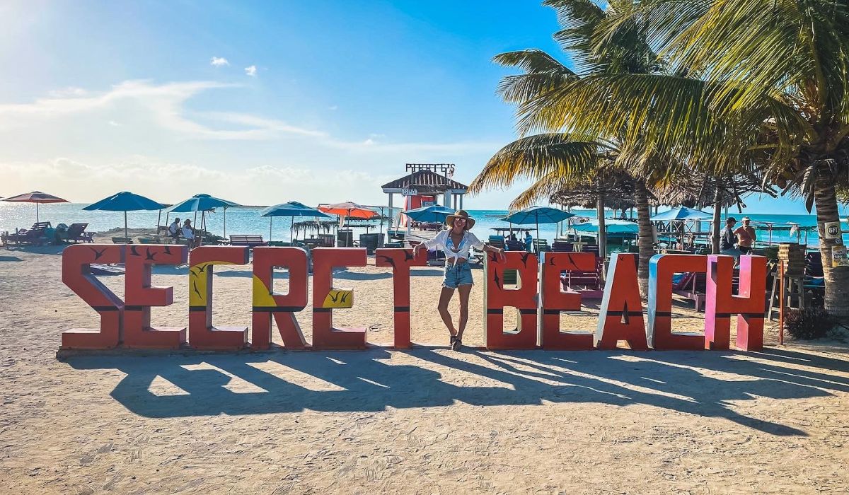 Kate poses in front of a large 'Secret Beach' sign at the entrance to Secret Beach in San Pedro, Belize. The beach behind her features colorful umbrellas, palm trees, and lounge chairs, with the ocean glistening under a clear blue sky.