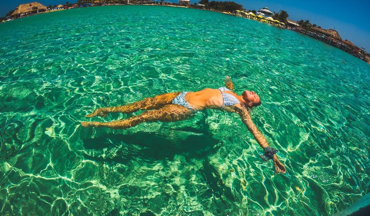 Kate floats peacefully in the crystal-clear turquoise waters at Secret Beach in San Pedro, Belize. She is wearing a bikini, with the sun shining down and the beach resort visible in the background.