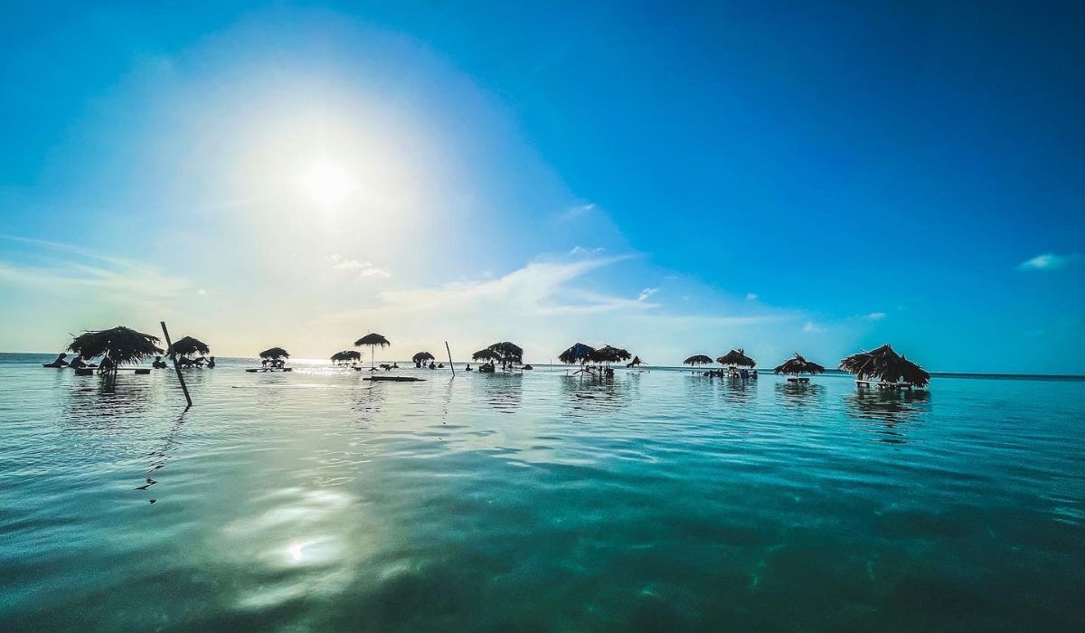 Thatched-roof huts stand in shallow, calm waters at Secret Beach in San Pedro, Belize, under a bright, sunny sky. The serene setting is enhanced by the clear blue water and the reflection of the sun on the surface.