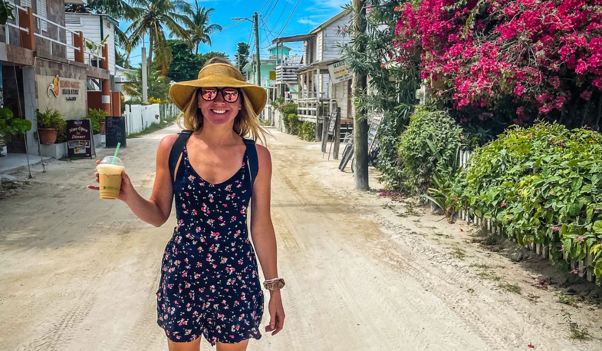 Kate enjoys a refreshing drink while strolling down a sandy street in Caye Caulker, Belize. She is wearing a floral dress, a wide-brimmed hat, and sunglasses, surrounded by colorful tropical flowers and laid-back island vibes, capturing the charm of this popular destination.