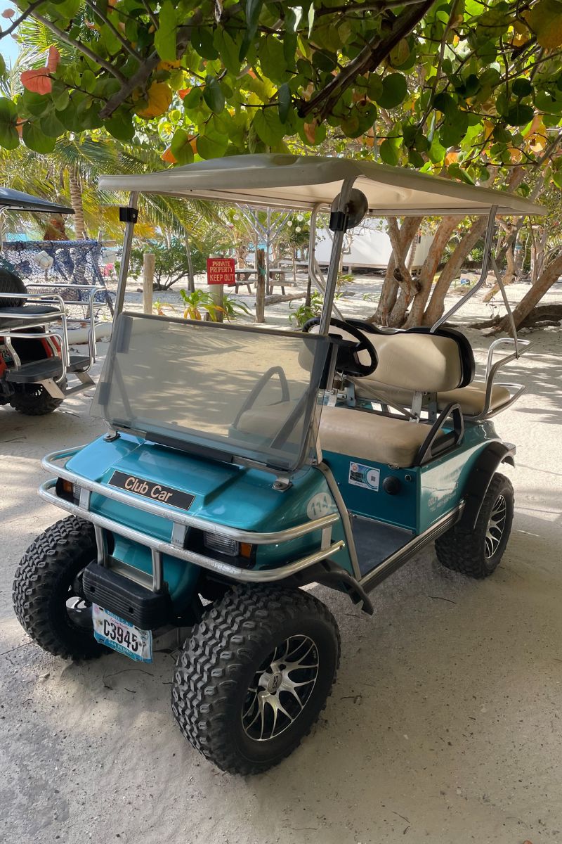 A turquoise golf cart parked on a sandy path in San Pedro, Belize, shaded by leafy trees. The cart features off-road tires and a beige canopy.