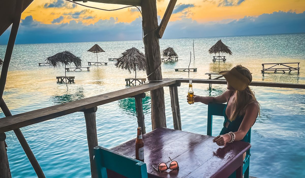 Kate enjoys a cold beer while watching the sunset from a beachfront bar at Secret Beach in San Pedro, Belize. The view includes thatched-roof huts in the shallow water, creating a tranquil and picturesque setting